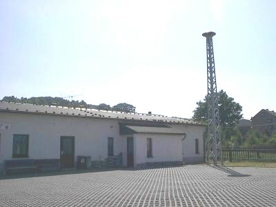 Gepflasterter Platz vor einem langen, weißen Flachbau mit Vordach; rechts daneben ragt ein Stahlgittermast mit Sirene in den hellen Himmel.
