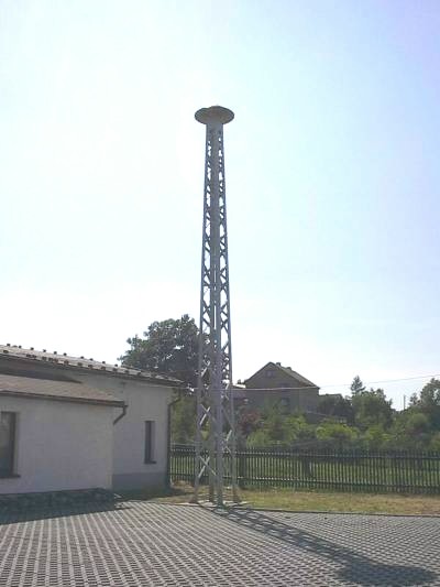 Hoher, schlanker Stahlgittermast mit einer Sirene an der Spitze neben einem weißen Gebäude, im Gegenlicht vor hellem Himmel aufgenommen.