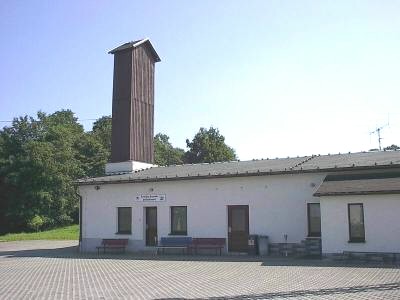 Weißes, eingeschossiges Gebäude mit flachem Dach und hohem, holzverkleidetem Schlauchturm; vor dem Eingang stehen Bänke auf einem gepflasterten Platz.