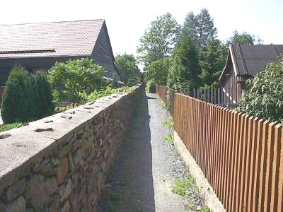 Schmaler Schotterweg verläuft zwischen einer Bruchsteinmauer links und einem braunen Holzlattenzaun rechts, dahinter Häuser und Bäume.