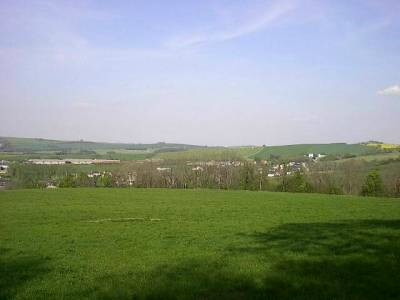 Weite Wiese im Vordergrund, dahinter eine Baumreihe und die verstreuten Häuser des Dorfes zwischen Feldern, darüber blauer Himmel mit Schleierwolken.