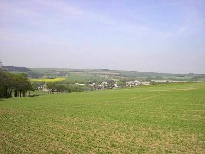 Junges grünes Getreidefeld am Hang, dahinter die Häuser des Dorfes, ein gelb blühendes Rapsfeld und sanfte Hügel am Horizont.