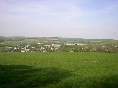 Blick über eine grüne Wiese auf das im Tal liegende Dorf mit hellen Häusern, dahinter Felder und bewaldete Höhenzüge unter leicht bewölktem Himmel.
