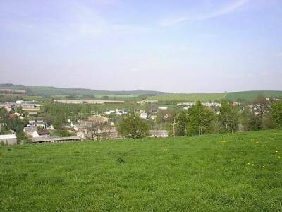 Blick über einen Wiesenhang auf Häuser und lange flache Stallgebäude am Ortsrand, rechts eine Baumgruppe, dahinter Felder und Höhenzüge.
