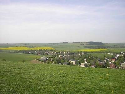 Blick von einem Wiesenhang auf Häuser mit dunklen Dächern zwischen Bäumen, dahinter zwei gelb blühende Rapsfelder und sanfte Hügel.