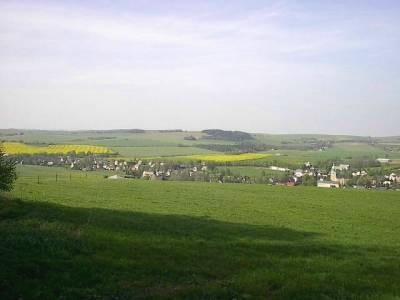 Weiter Blick über eine grüne, teils beschattete Wiese auf das Dorf im Tal, dahinter leuchtend gelbe Rapsfelder und bewaldete Hügel.