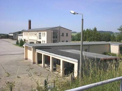Betriebsgelände mit zweigeschossigem, hellem Gebäude mit Schornstein, davor ein Flachbau mit offenen Unterständen; vorn eine Laterne und hohes Gras.
