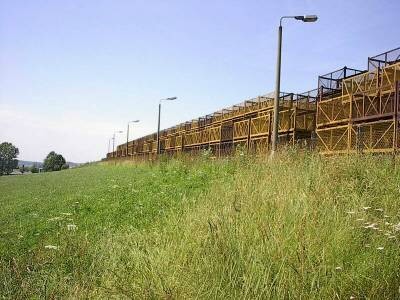Lange Reihe hoch gestapelter, gelber Gitterkisten hinter einer Wiese mit hohem Gras, davor mehrere Laternenmasten unter blauem Himmel.
