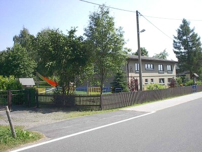 Zweigeschossiges graues Gebäude hinter einem Holzzaun an der Straße, davor ein Garten mit hohen Bäumen und einer roten Rutsche.