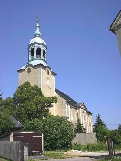 Gelb verputzter Kirchturm mit Uhr, heller Haube und Laterne vor tiefblauem Himmel; davor Bäume, eine Mauer und eine Garage mit braunem Tor.