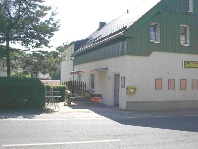 Haus an der Straße mit weißem Erdgeschoss und grün verkleidetem Obergeschoss, gelbem Briefkasten neben dem Eingang und Markise über einer Sitzecke.