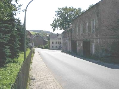 Leere Dorfstraße mit gepflastertem Gehweg und Zaun links, rechts ein großes altes Steingebäude; im Hintergrund Häuser und ein bewaldeter Hügel.