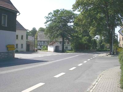 Leere Straße mit Mittelstreifen führt auf einen Platz mit großen Laubbäumen, dahinter Fachwerkhäuser und ein kleines weißes Häuschen.