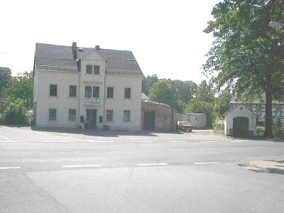 Helles dreigeschossiges Haus mit Mittelgiebel an einer weiten Straßenkreuzung, rechts ein großer Baum, ein kleines weißes Häuschen und ein rotes Auto.
