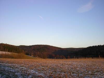 Leicht verschneites Stoppelfeld im rötlichen Abendlicht, dahinter bewaldete Hügel unter klarem, blauem Himmel.