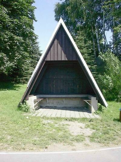 Dreieckige, vorn offene Schutzhütte aus dunklem Holz mit Sitzbänken auf einer Wiese am Rand der Bahn, dahinter Bäume.