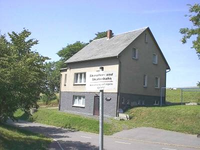 Zweigeschossiges Haus mit Schieferdach auf einer Wiese; davor ein weißes Schild mit der Aufschrift „Skiroller- und Skaterbahn“.
