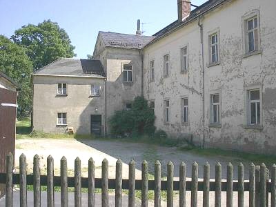 Großes, mehrgeschossiges Gebäude mit stark abblätterndem Putz an einem leeren Hof, im Vordergrund ein Holzlattenzaun.