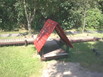 Niedriger Holzsteg auf einer Wiese mit einem kleinen roten Satteldach in der Mitte, dahinter Bäume und Sträucher.
