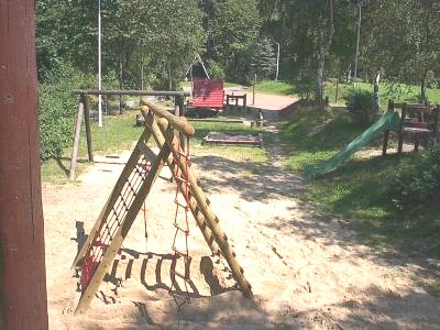 Hölzernes Klettergerüst mit rotem Netz und Strickleiter auf einem sandigen Spielplatz, dahinter eine grüne Rutsche, Bänke und Bäume.