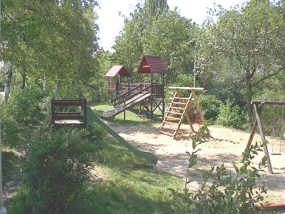 Spielplatz unter Bäumen mit hölzernem Kletterturm mit roten Dächern, grüner Rutsche, Kletternetz und Schaukel auf Sandboden.