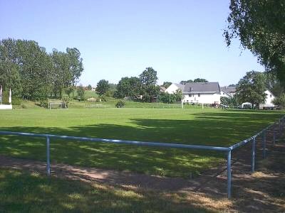 Fußballplatz mit Tor hinter einem Metallgeländer, im Hintergrund Bäume und ein weißes Wohnhaus unter blauem Himmel.