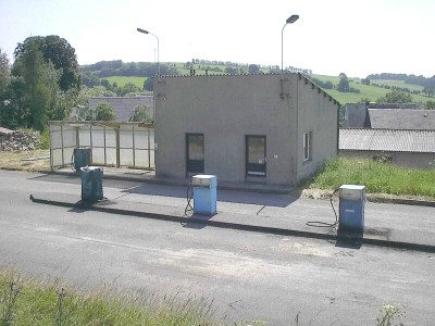 Drei alte Zapfsäulen auf einer Betoninsel vor einem grauen Flachbau; im Hintergrund Dächer, Felder und Hügel.