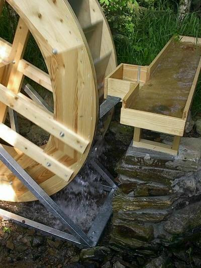 Neues hölzernes Wasserrad in einem Metallgestell; daneben führt eine Holzrinne auf einem Bruchsteinsockel das Wasser heran.