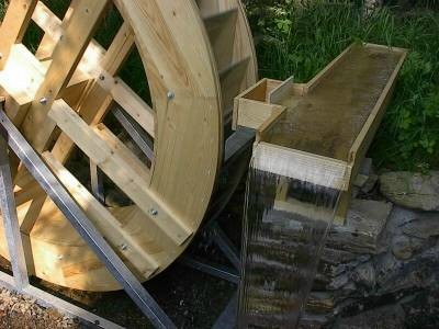 Aus einer Holzrinne fällt Wasser in einem breiten Schleier neben dem hölzernen Wasserrad herab, dahinter grünes Ufergebüsch.
