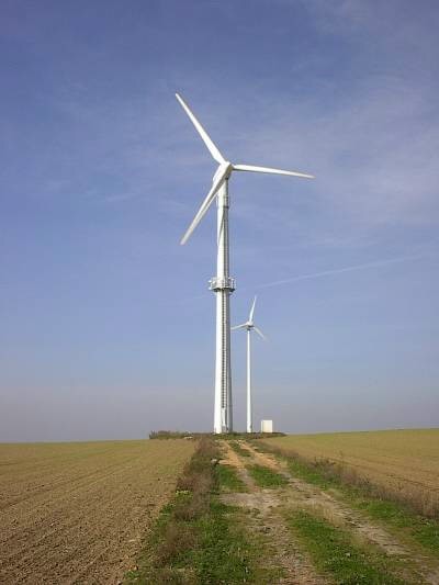 Zwei weiße Windkraftanlagen hintereinander am Ende eines Feldwegs zwischen abgeernteten Äckern, unter blauem Himmel mit Schleierwolken.
