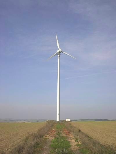 Einzelne weiße Windkraftanlage mit drei Rotorblättern am Ende eines grasbewachsenen Feldwegs zwischen Äckern, darüber blauer Himmel.