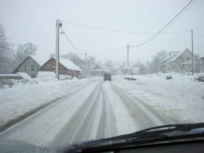 Blick durch die Windschutzscheibe auf eine schneebedeckte Straße mit Fahrspuren, voraus ein Auto, am Rand Häuser und Strommasten im Schneetreiben.