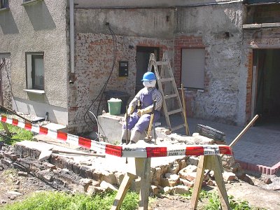 Eine Puppe mit blauem Bauhelm sitzt vor einer unverputzten Hauswand neben einer Holzleiter, davor Bruchsteine und rot-weißes Absperrband.