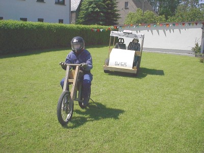 Auf einer Wiese sitzt eine Puppe mit Helm auf einem aus Holz gebauten Motorrad, dahinter zwei Puppen mit Helm in einem Holzauto mit dem Kennzeichen „GW 625“.