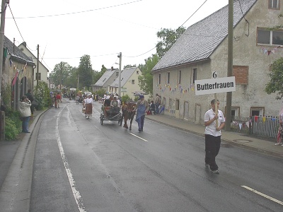 Eine Frau trägt das Schild „6 Butterfrauen“ über die nasse Dorfstraße; hinter ihr folgen eine Pferdekutsche und Frauen in historischen Kleidern.