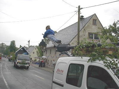 Auf dem Dach eines weißen Transporters ist eine Straßenlaterne mit einem Mast montiert, an dem eine Puppe in blauer Arbeitskleidung mit Steigeisen hängt.