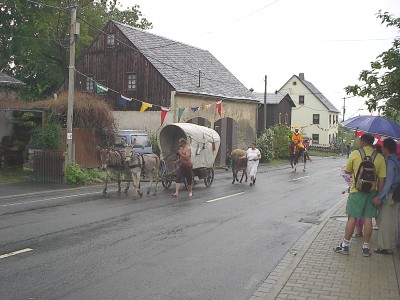 Zwei Esel ziehen einen kleinen Planwagen über die nasse Dorfstraße, begleitet von mehreren Personen und einem weiteren Tier; rechts Zuschauer unter Regenschirmen.