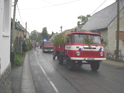 Zwei rot-weiße Feuerwehrfahrzeuge fahren hintereinander über die nasse Dorfstraße; das vordere, ein älterer Lastwagen, ist mit Birkenzweigen geschmückt.
