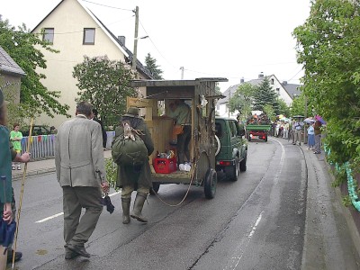 Zwei Männer in grüner Jagdkleidung, einer mit Rucksack, gehen hinter einem Geländewagen mit einem hölzernen Hochsitz auf dem Anhänger her.