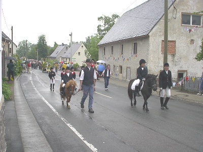 Kinder in Reitkleidung reiten auf Ponys über die nasse Dorfstraße, begleitet von Erwachsenen zu Fuß; am Straßenrand stehen Zuschauer mit Regenschirmen.