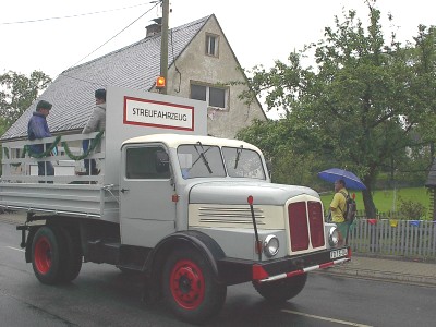 Alter grau-weißer Lastwagen mit roten Felgen und dem Schild „Streufahrzeug“; auf der Ladefläche stehen zwei Männer, am Straßenrand ein Zuschauer mit Regenschirm.