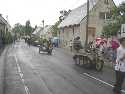 Ein historisches Kettenkrad mit zwei Personen in Uniform fährt über die nasse Dorfstraße, dahinter folgen ein Motorrad mit Beiwagen und weitere Militärfahrzeuge.