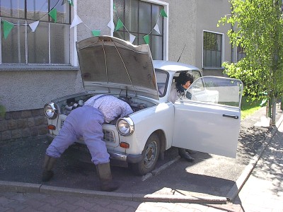 Eine Puppe im blauen Arbeitsanzug beugt sich unter die offene Motorhaube eines weißen Trabants, eine zweite Figur steht an der geöffneten Fahrertür.