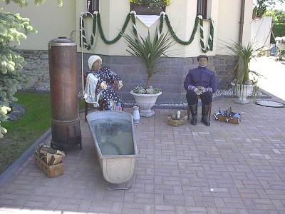 Vor einem geschmückten Haus stehen ein alter Badeofen und eine Zinkbadewanne; dahinter sitzen zwei Puppen, eine Frau im Kittel und ein Mann in Arbeitskleidung.