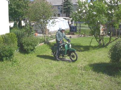 Eine Puppe in grüner Uniform mit Helm sitzt auf einem grünen Moped, das auf einer Wiese zwischen Sträuchern und Obstbäumen steht.
