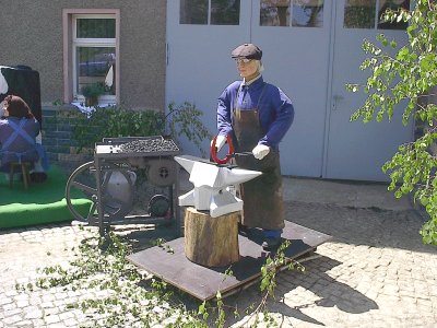 Eine Puppe mit Schiebermütze und Lederschürze steht mit einem Hufeisen an einem Amboss auf einem Holzklotz, daneben eine fahrbare Feldschmiede mit Kohle.
