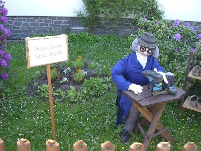 Eine Puppe mit Hut, Brille und weißem Bart sitzt im Garten an einem Holztisch mit Stiefeln; daneben ein Schild „Schuhladen Kurt Bach“ und ein Regal mit Schuhen.