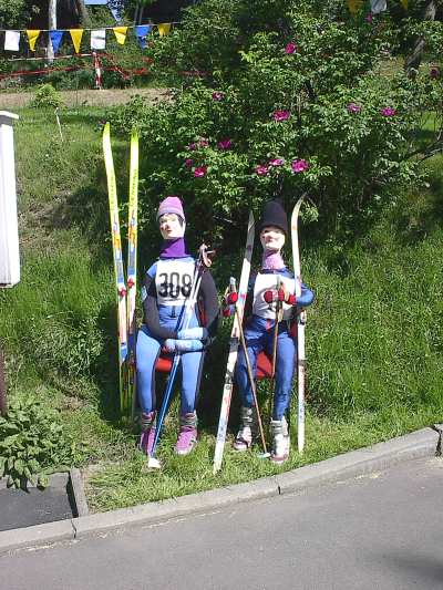 Zwei Puppen in Langlaufanzügen mit Mützen sitzen am Straßenrand vor einem blühenden Strauch, eine trägt die Startnummer 308; neben ihnen stehen Skier und Stöcke.