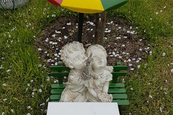 Helle Gartenfigur zweier Kinder auf einer kleinen grünen Bank: Der Junge zeigt mit dem Finger nach oben, über ihnen ein bunter Schirm, ringsum Blütenblätter im Gras.