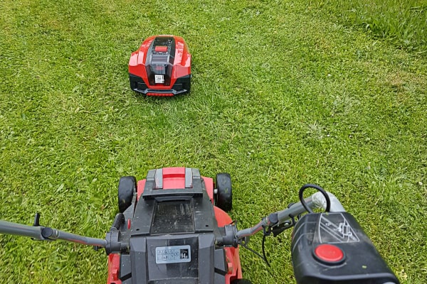 Blick vom Holm eines roten Rasenmähers über den Rasen auf einen kleinen roten Mähroboter, der ein Stück voraus im Gras fährt.